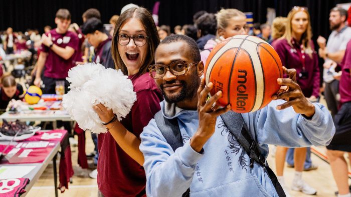 Two students enjoying a uni sports fair, one is holding a basketball and the other is holding a cheerleading pompom.