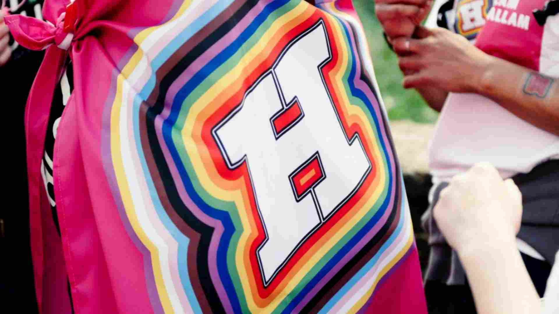 A photo of a flag / cape with the Hallam H with the pride rainbow colours around the edge of it and people in the background celebrating with the Hallam logos and rainbow colours. 